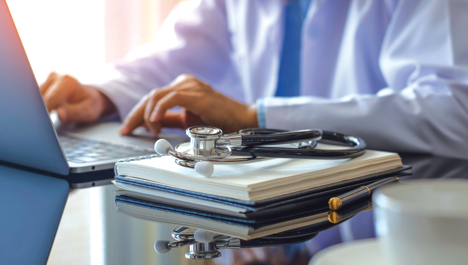 photo of a medical professional typing on a laptop with a notebook and stethoscope sitting on a table in the foreground