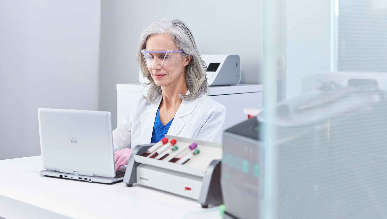 Woman in lab coat sitting at desk typing on computer with laboratory equipment in the foreground