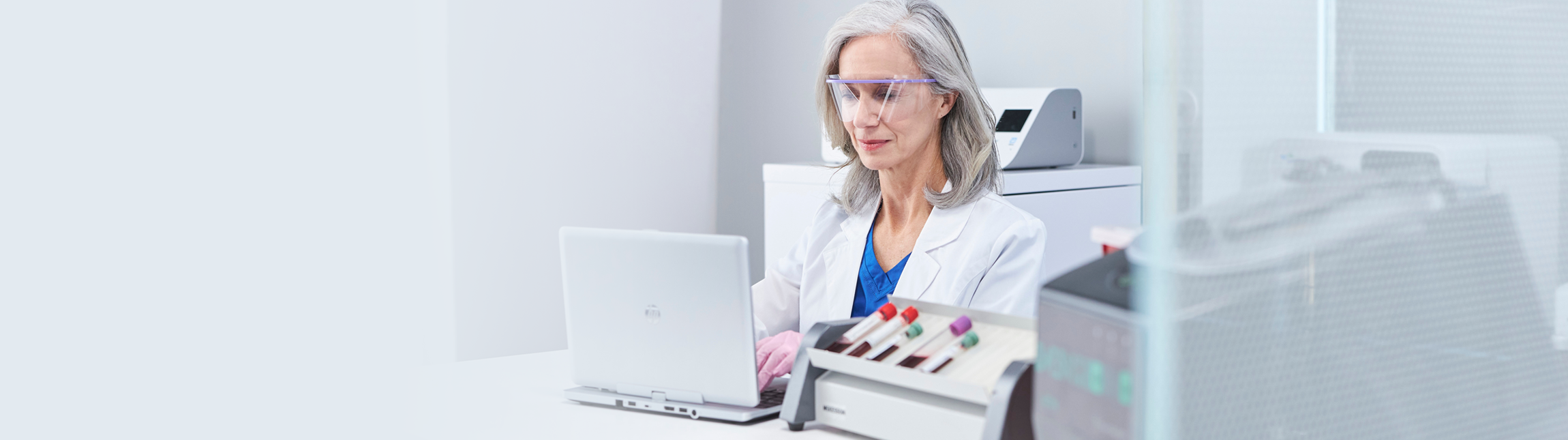 Woman in white lab coat typing on laptop with safety glasses and gloves on and lab equipment present in the foreground