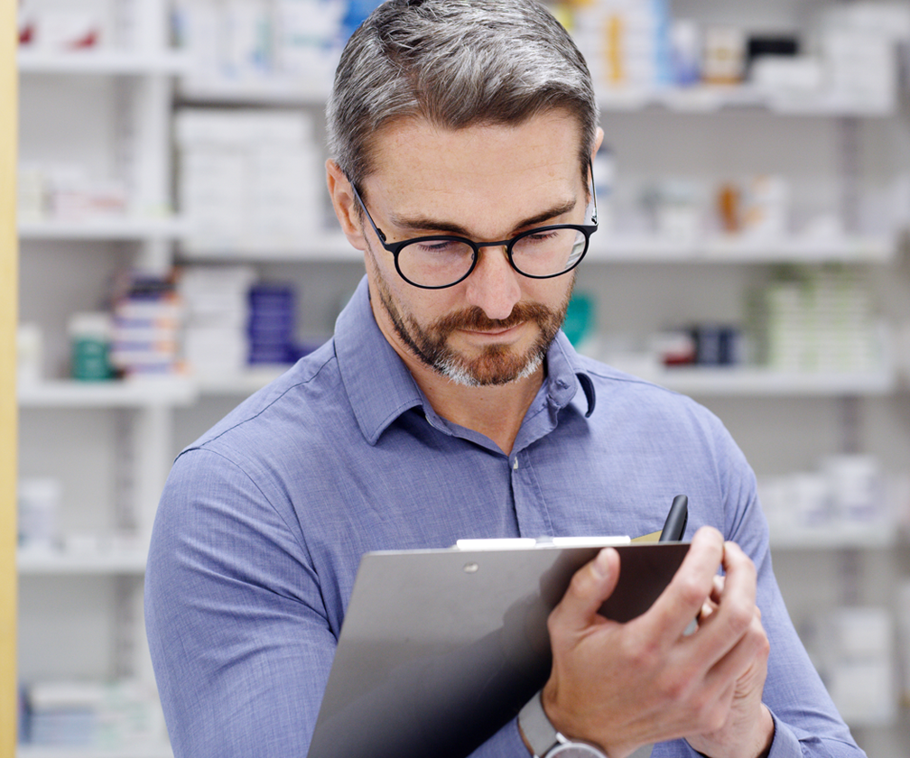 photo of male pharmacist making notations on a clipboard in an inventory setting with pharmaceuticals products on shelves behind him