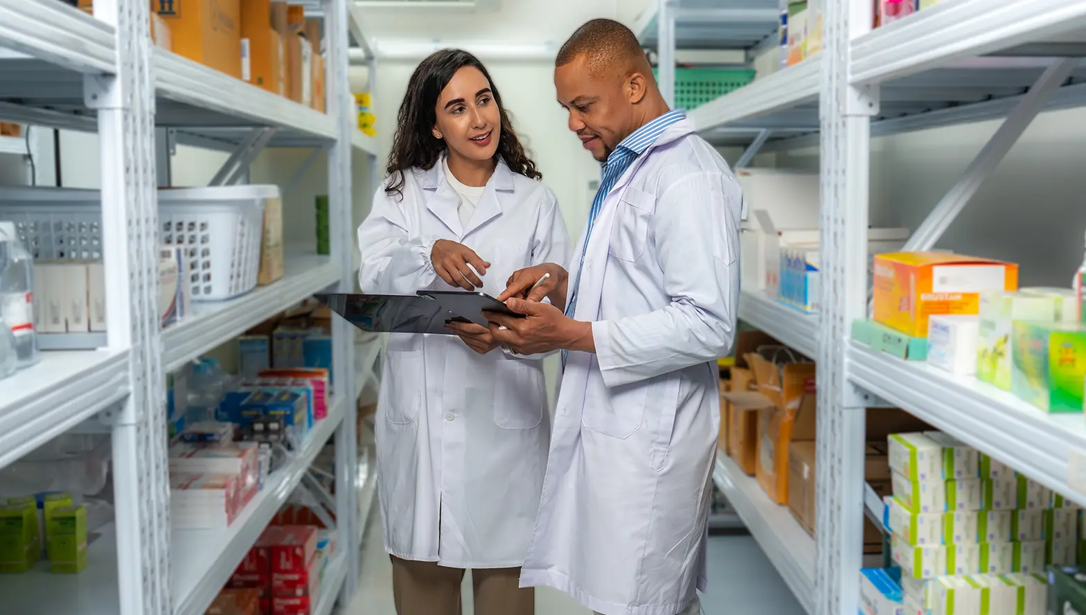 Two people in lab coats review a tablet while standing in a well-organized medical supply storage aisle.