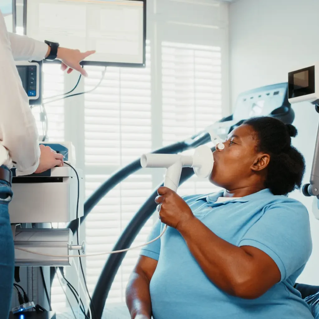 photo of a female patient breathing into a spirometer in a clinic setting as a medical professional offscreen reads the results