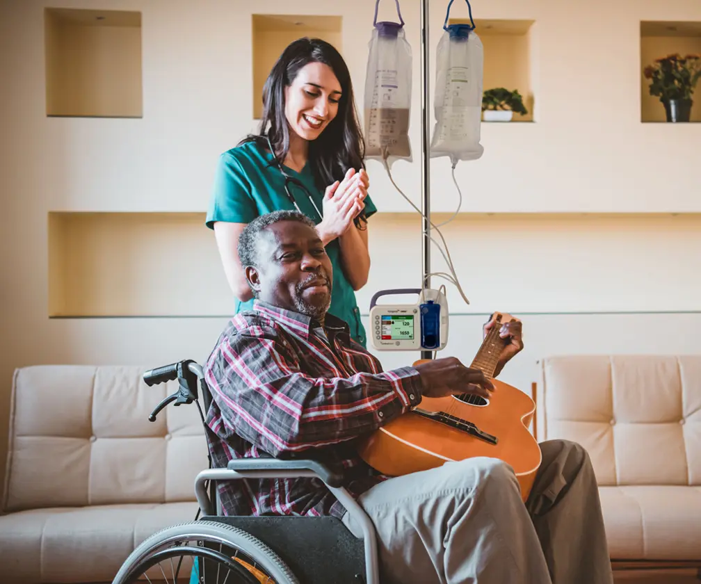 photo of a female nurse clapping as she listens to an older male patient in a wheelchair with IV and enteral feeding bag strum a guitar in a healthcare setting