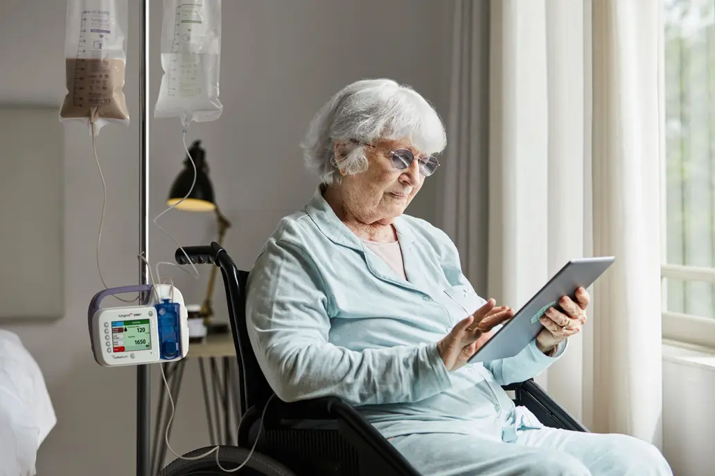 photo of an older female patient receiving an enteral pump feeding while sitting in a wheelchair in a healthcare setting